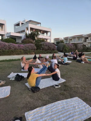 Young participants practising partner yoga and mindfulness exercises outdoors on grass with seaside residences in the background.
